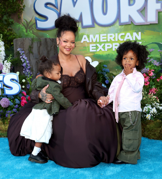 Rihanna poses at a premiere with her sons Riot and RZA. She wears her hair in a high bun and wears a brown dress and...