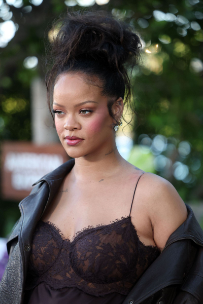 Rihanna poses at a premiere in a high messy bun. She wears a brown lace gown and leather jacket.