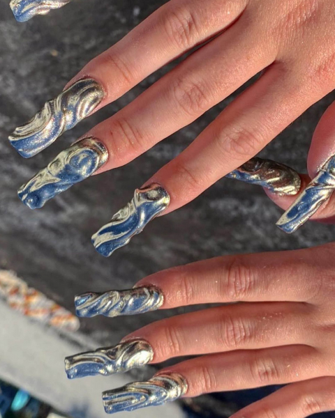 Hands facedown and flat showcasing very long wavy and metallic nails.