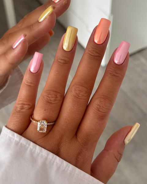 Hand coming out of a white sleeve with a ring and alternating pink yellow and orange metallic nails.